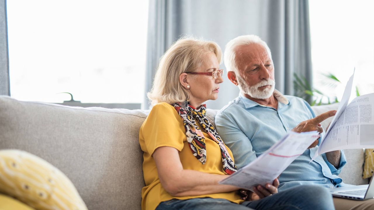 A retired couple sitting on their couch look through a pile of bills.