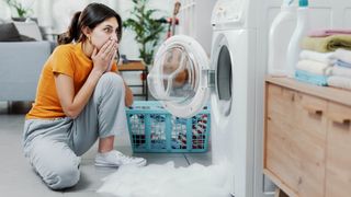 An open washing machine with suds on the floor and a woman looking concerned