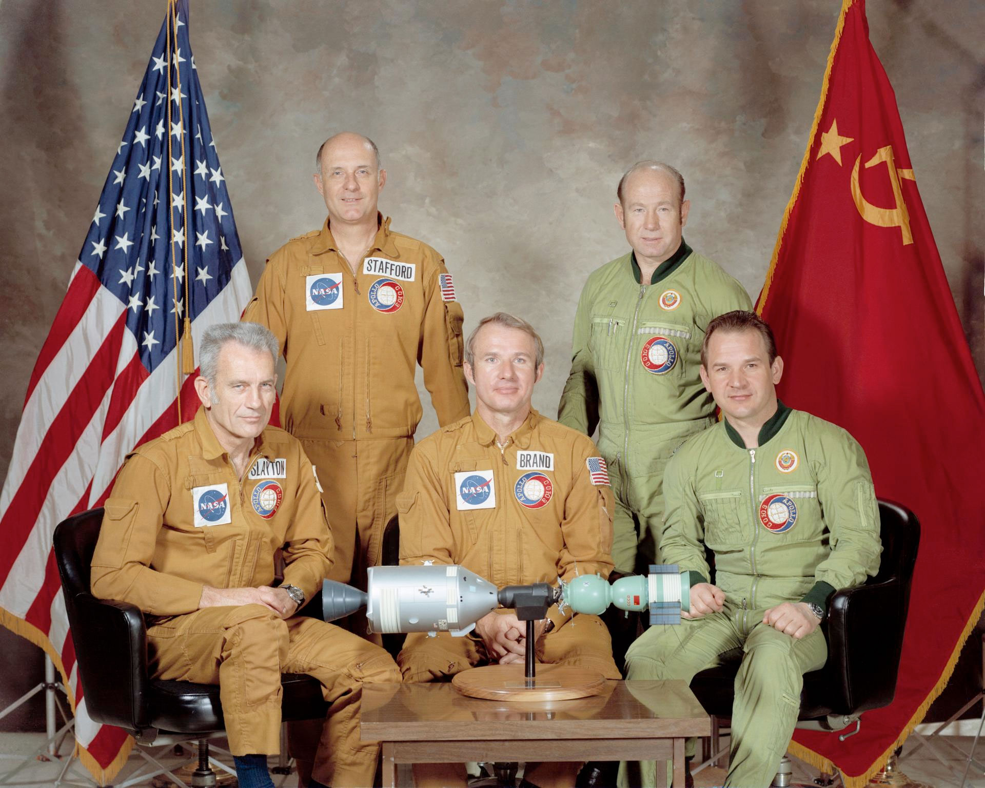 five men, three in orange-tan flight suits and two in olive green flight suits, pose together for a portrait. They are flanked by flags of the United States and the Soviet Union.