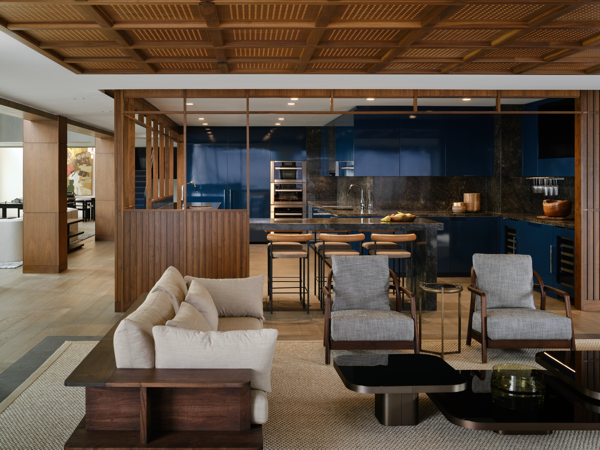 open plan space with living room with beige timber sofa and two gray armchairs, and a high-gloss black glass coffee table. in the background is a glossy blue kitchen with timber elements, including a lattice-style timber coffered ceiling
