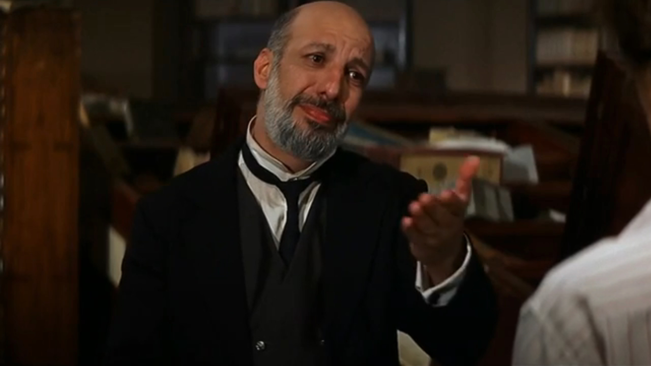 Erick Avari gestures expressively as he stands in a wrecked library in The Mummy.