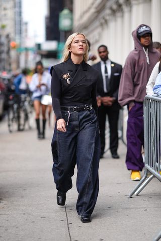 Savannah White wears silver rimmed sunglasses, silver earrings, black long sleeve shirt, black criss cross blazer tailored vest with sun and moon brooches, black leather belt, dark blue denim jean baggy pants, shiny boots leather shoes, outside Who Decides War, during the New York Fashion week Spring/Summer 2025 on September 07, 2024 in New York, New York.