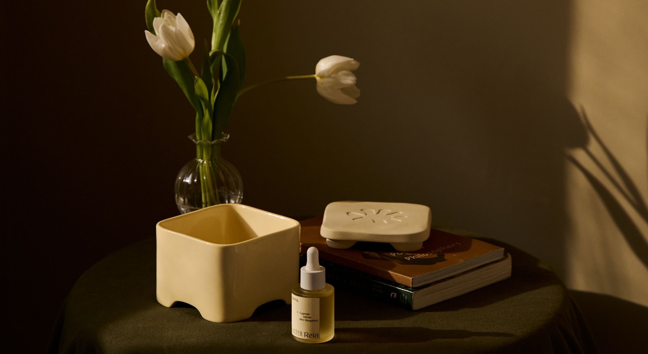 A room filled with light of a sunset boasts a round table topped by a fragrance diffuser in the shape of a cream box with a floral motif on its top, two books, and a glass vase filled with flowers.
