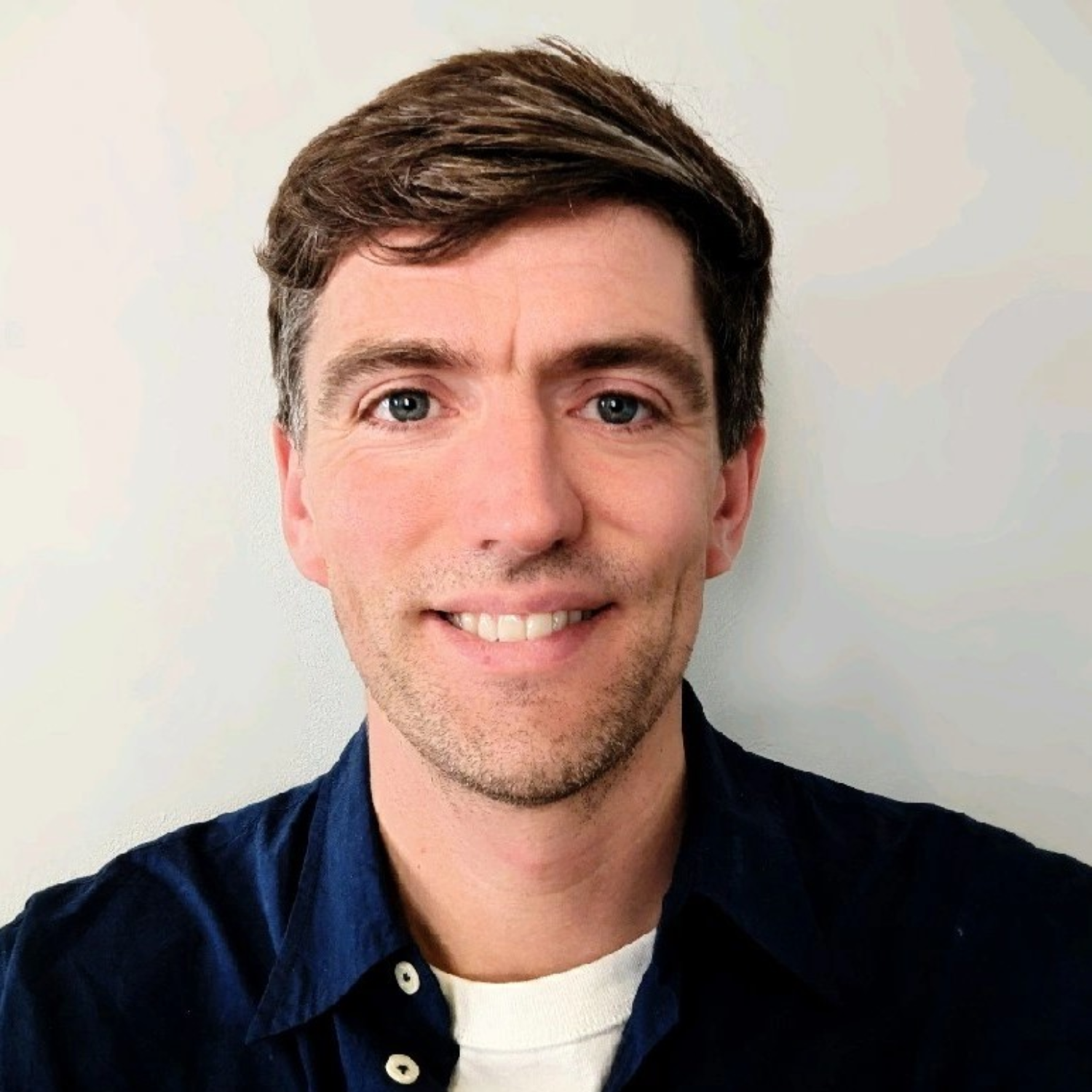 A headshot of a man in a navy button down with a white round-neck t-shirt underneath