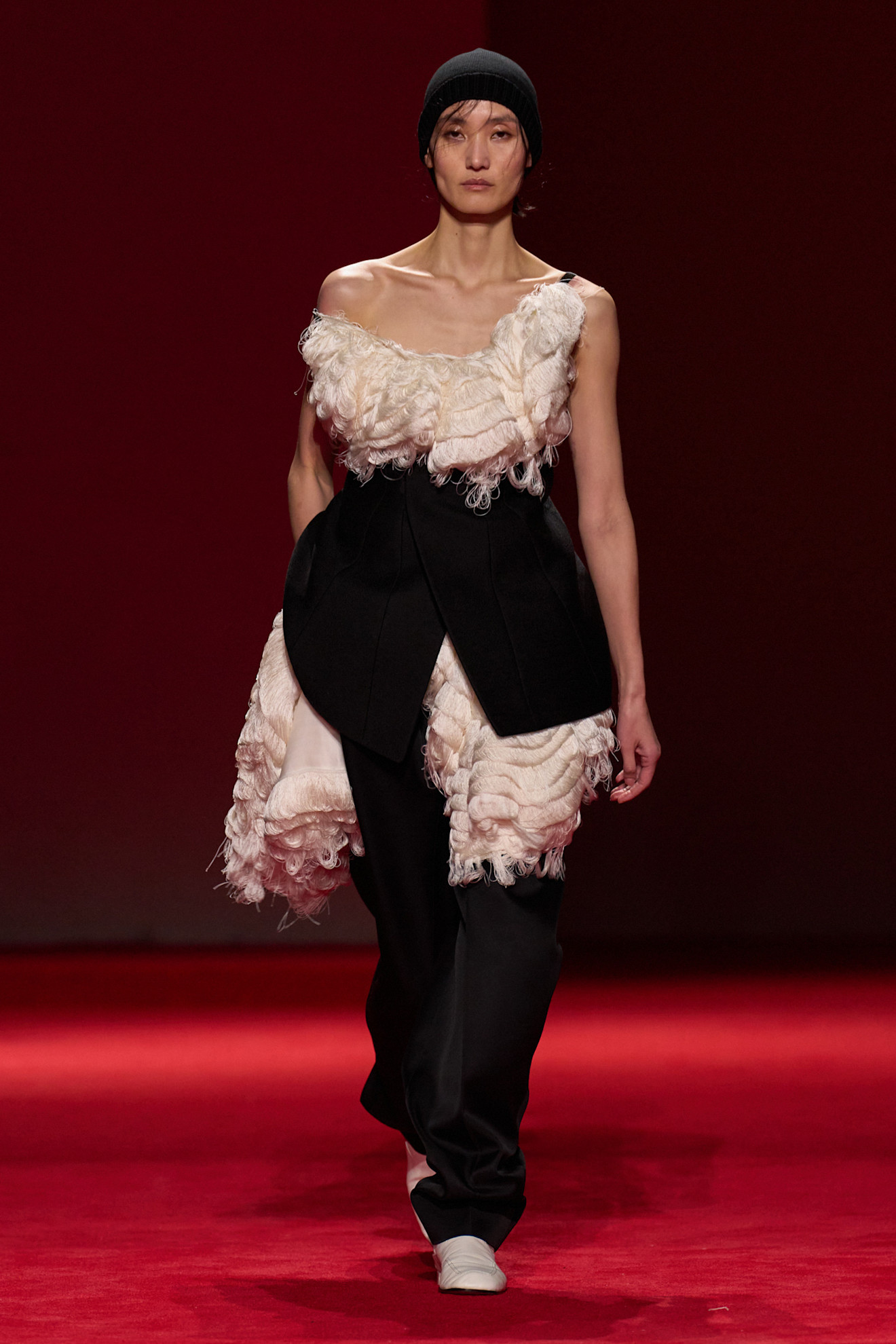 A model walking in the F/W 26 Bottega Veneta show during Milan Fashion Week wearing a black beanie, fringe cream top, and black pants.