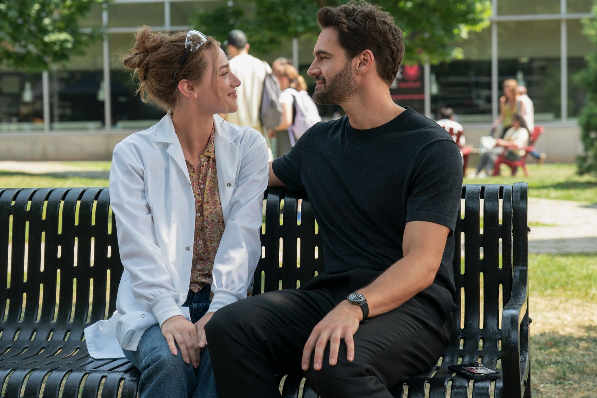 A woman in a lab coat (Lili Reinhart as Olive Smith) and a man in all-black (Tom Bateman as Dr. Adam Carlsen) sit on a park bench, in a still from 'The Love Hypothesis.'