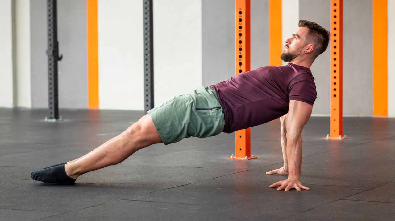 A man performs a reverse plank in an empty room, with metal beams behind him. He wears a t-shirt and shorts. He is on the ground, with his back facing the floor and his body held aloft in one straight line. His feet and hands are on the floor and his arms are straight.