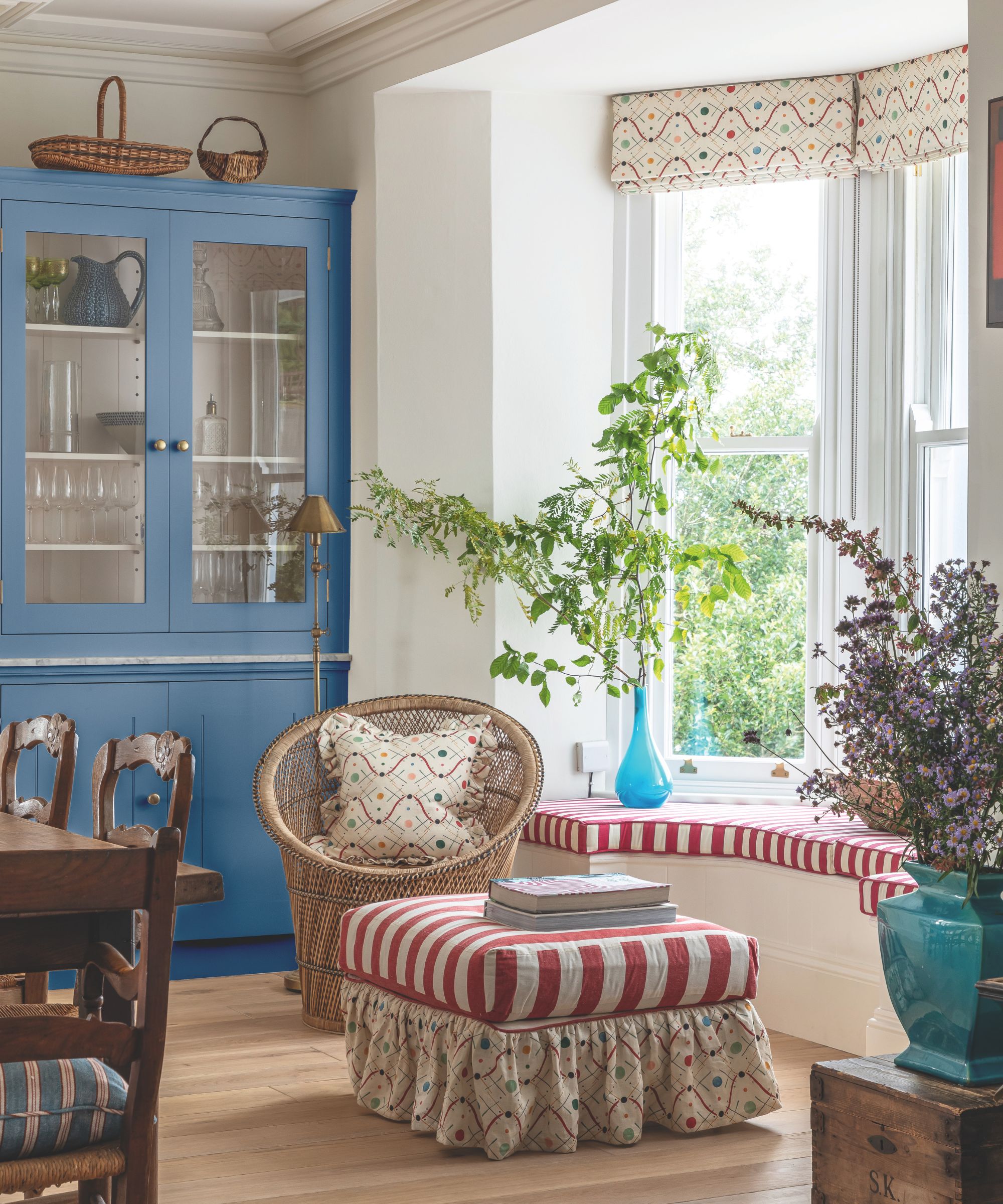 A sunlit dining room designed by Sarah Southwell, featuring a striking cornflower blue hutch and a bay window seat with red-and-white striped cushions. The room is styled with a vintage wicker peacock chair, a ruffled ottoman, and vibrant floral arrangements, creating a playful, English country aesthetic.