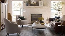 White living room with brick walls. There's two gray armchairs on the left hand side and a leather couch on the right. In between is an oval upholstered ottoman