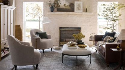 White living room with brick walls. There's two gray armchairs on the left hand side and a leather couch on the right. In between is an oval upholstered ottoman
