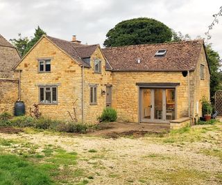 A stone cottage with two storeys and a pitched roof with ground being dug in preparation for a glass box extension