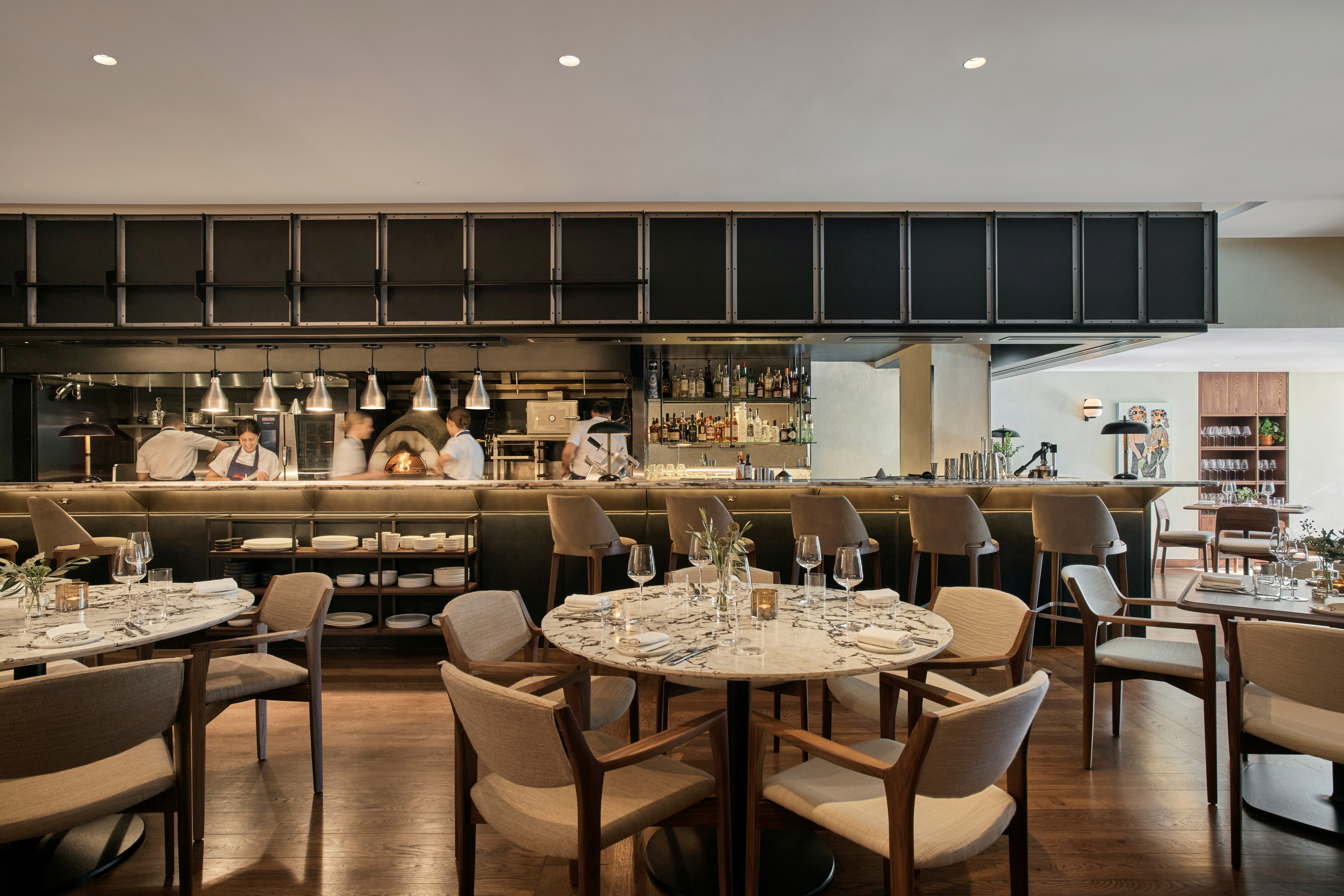 An open kitchen sitting in the middle of a minimalist-designed restaurant with white marble and wood tables and white shearling upholstered wooden chairs shows a brigade at work behind its marble, steel, and iron counter.