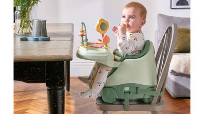 A baby holds a piece of food to its mouth whilst sitting in the Mamas and Papas Bug 3-in-1 Floor & Booster Seat with Activity Tray near a dining table
