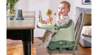 A baby holds a piece of food to its mouth whilst sitting in the Mamas and Papas Bug 3-in-1 Floor &amp; Booster Seat with Activity Tray near a dining table