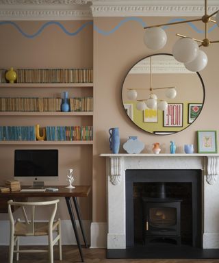 living room with pink walls, fireplace with round mirror, workspace in alcove with shelving