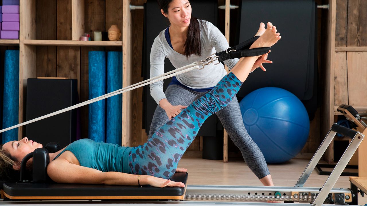 Woman lying on her back on a reformer machine, with her legs extended and raised, and her feet in straps attached to ropes which are attached behind the woman&#039;s head. An instructor stands over her adjusting the position of the other woman&#039;s legs with her hands.