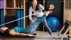 Woman lying on her back on a reformer machine, with her legs extended and raised, and her feet in straps attached to ropes which are attached behind the woman's head. An instructor stands over her adjusting the position of the other woman's legs with her hands.