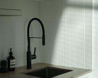 Kitchen faucet in matte black, pictured in shadow against white ridged vertical splashback tiles and steel sink area