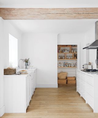 Minimalist white kitchen with wood floor, pantry in background with wooden shelves and baskets