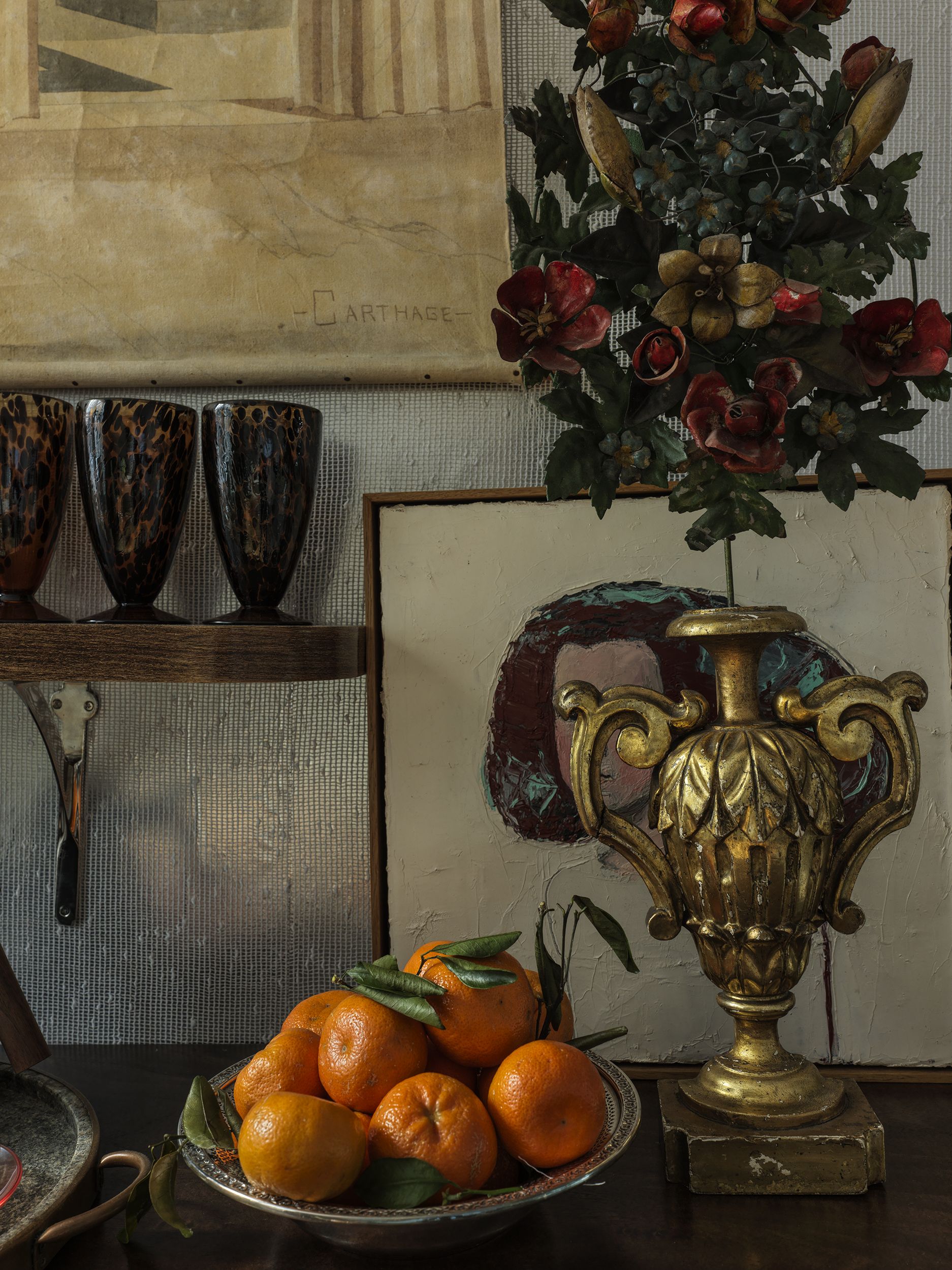 A close-up of a tabletop with a gold vase and a plate of tangerines, backed by silver wallpaper. 