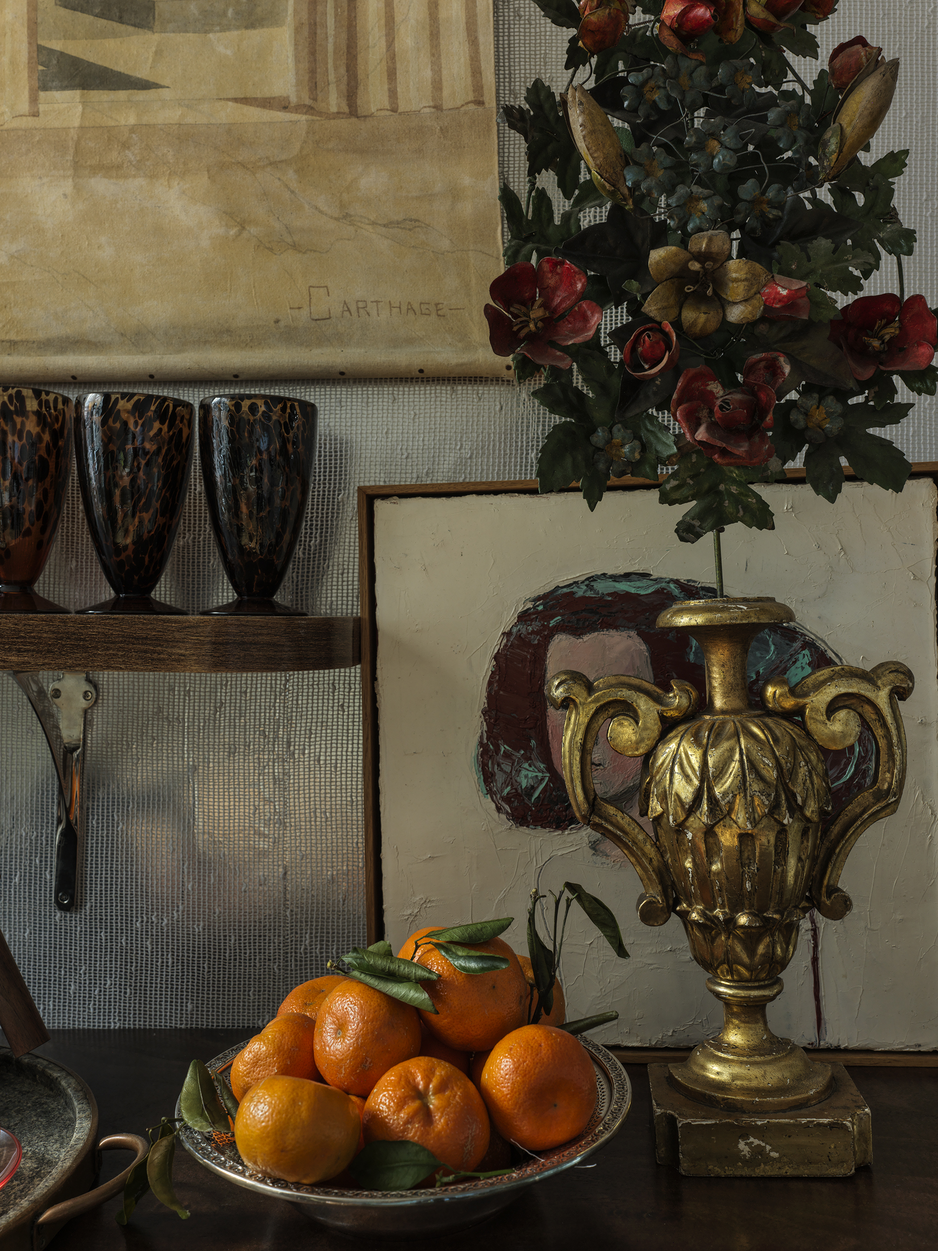 A close-up of a tabletop with a gold vase and a plate of tangerines, backed by silver wallpaper. 