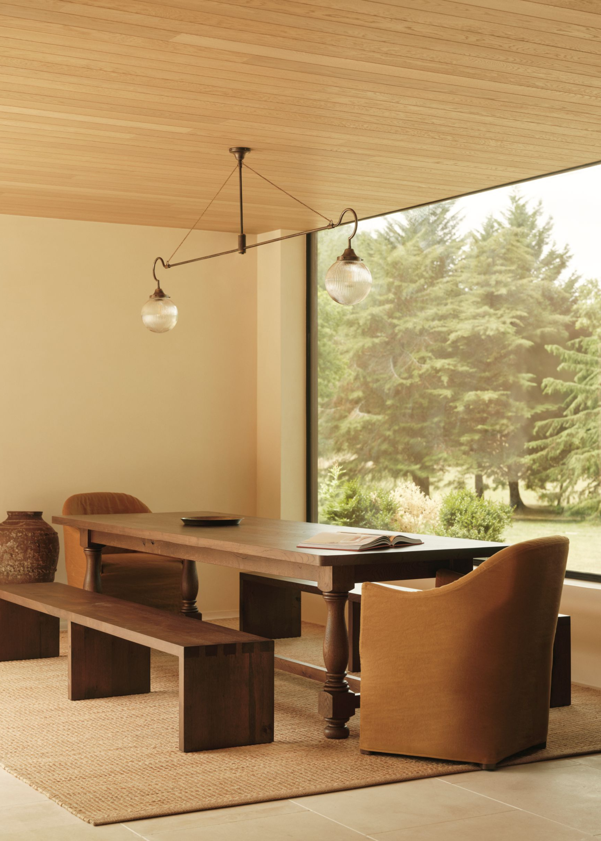 a dining area with a large, dark wooden table with matching benches, with a large floor to ceiling window opposite with views of nature