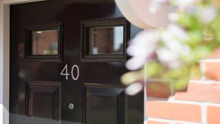 Black front door with silver house number and hanging basket by brick wall