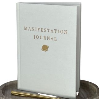 A white manifestation journal on a wooden bowl with a gold pen