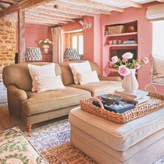 living room in small Cotswold cottage with pink painted walls, upholstered footstool and olive green sofa, rugs on floor, wicker tray, beam ceiling