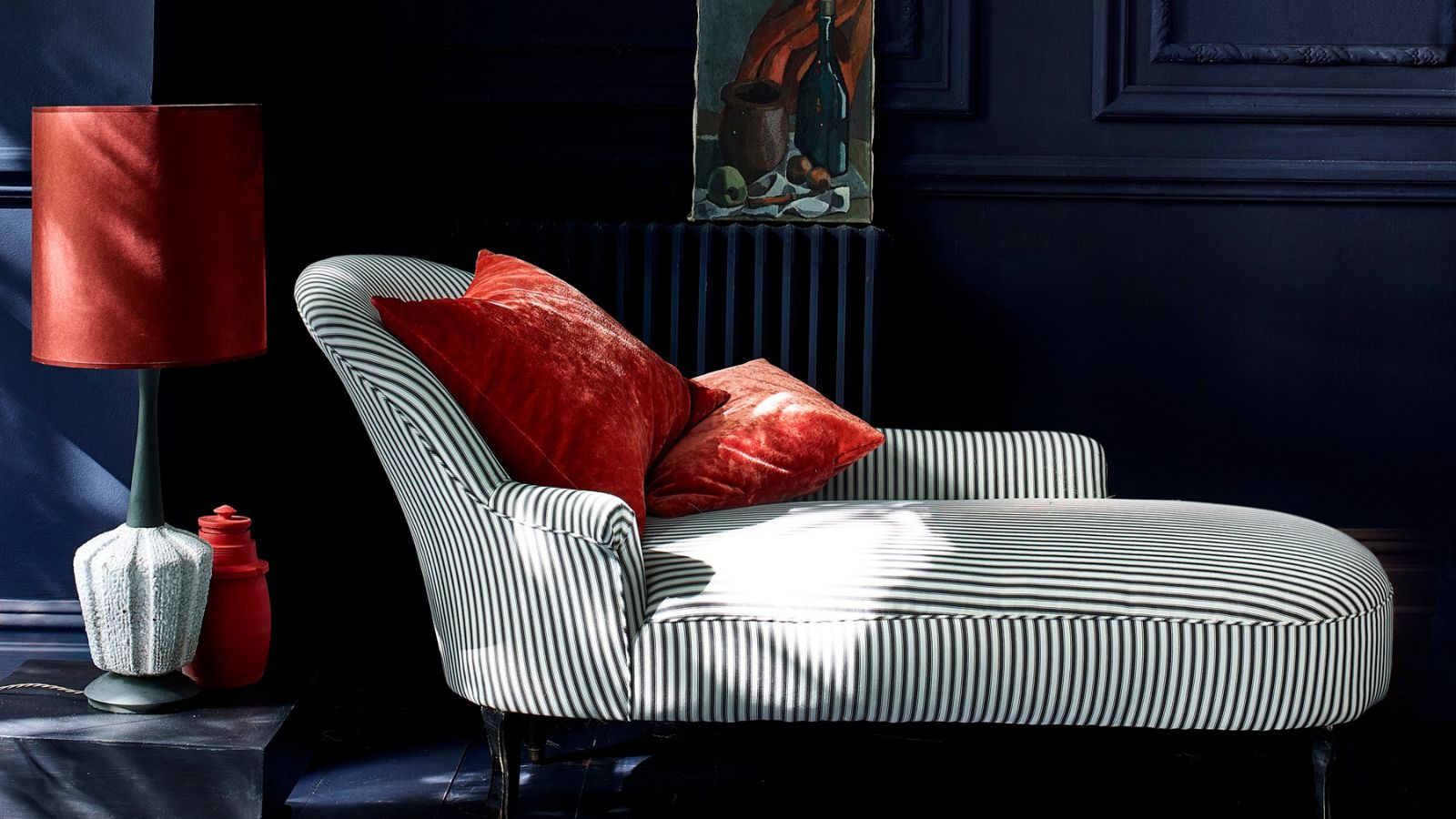 A dramatic dark navy panelled room with a monochrome striped chaise longue, terracotta velvet cushions, a rust red lamp and a small oil painting propped against a painted radiator.