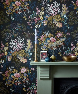 Close up of dark botanical wallpaper near painted green fireplace, mantel decorated with candles, decorative ornaments, flowers