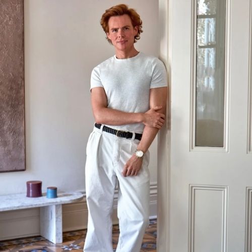 Image of a person in a white T-shirt and white pants with a black belt leaning against a white doorway.