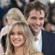 Robert Pattinson laughs and sticks his tongue out while wearing a white shirt and black suit on the Met Gala red carpet, and partner Suki Waterhouse smiles with long wavy golden blonde hair