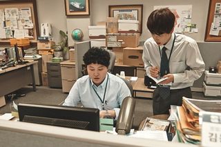 a man sits at a computer desk as another man stands behind him and takes notes, in the workplace k-drama 'misaeng incomplete life'