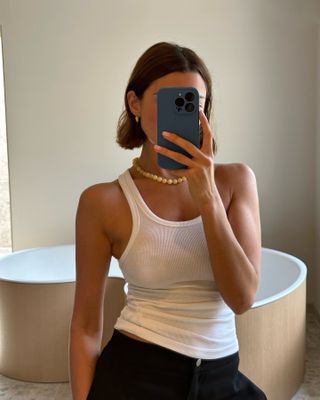 Woman taking a mirror selfie wearing a white tank top.