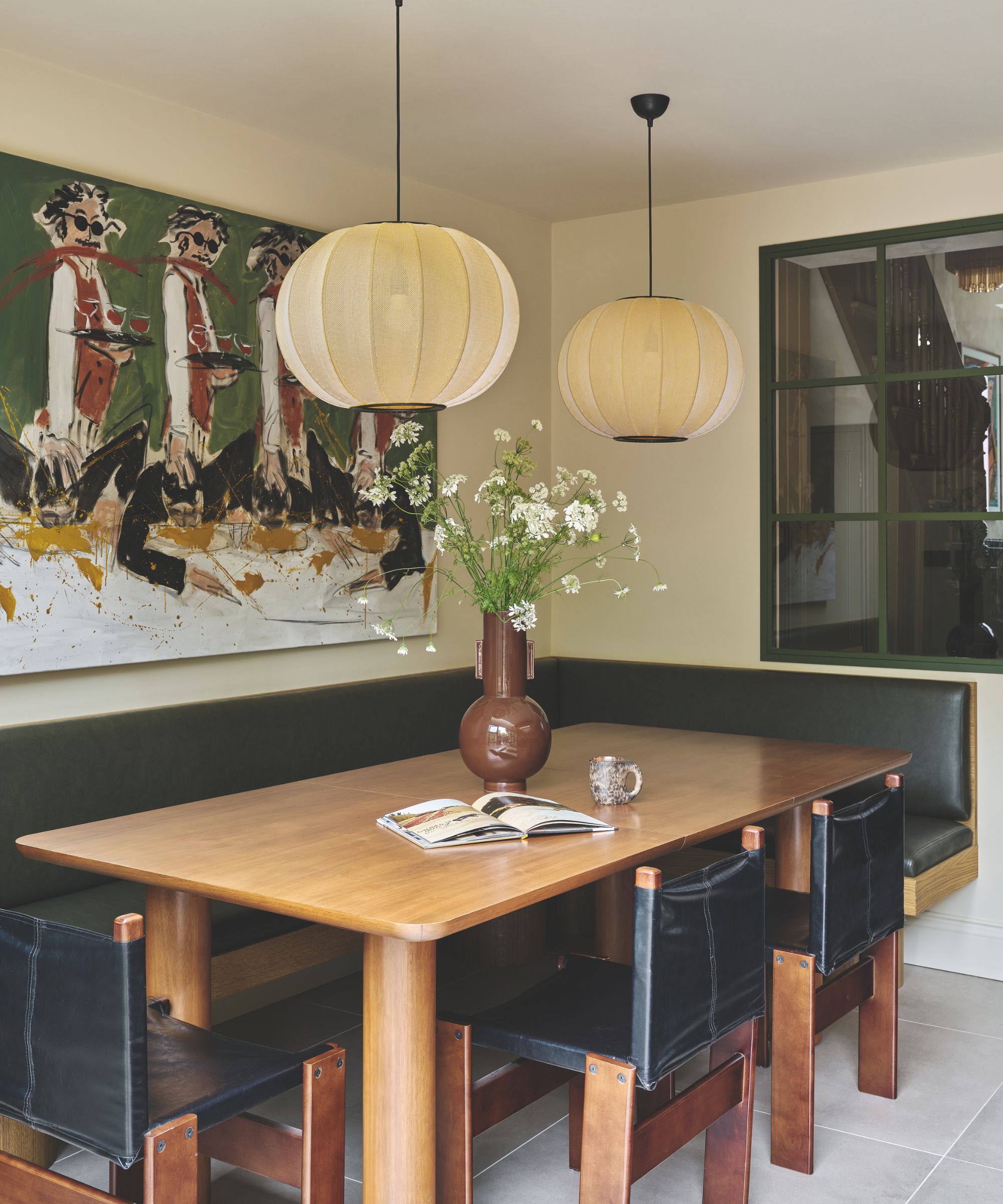 Dining nook with wooden rectangular dining table, leather and wooden dining chairs, leather banquette, two lantern pendant lights above the table, and an abstract painting of waiters holding wine