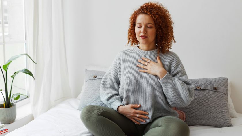 a woman sat cross legged on a bed inhaling with her eyes closed and hands over her chest and stomach