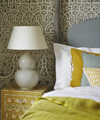 A close up of a gray headboard, white pillows and a gray and white lamp on a bohemian bedside tablee