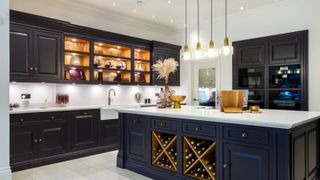 A sophisticated kitchen with deep navy in-frame cabinets and a large central island. Glass-fronted cupboards glow with warm lighting. Brass handles, pendant lights and a built-in wine rack add elegance to the contemporary, high-end design.