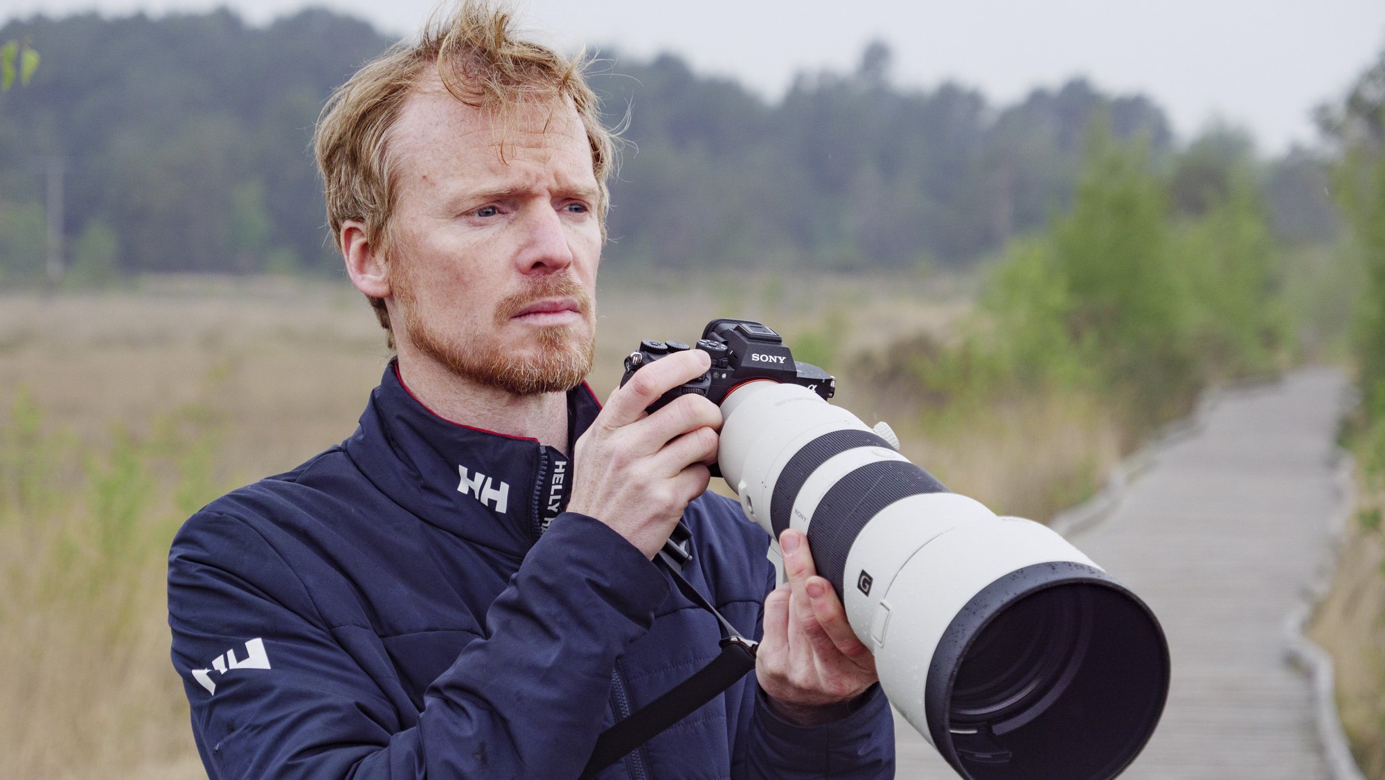 Man holding the Sony A7R VI camera with FE 400-800mm F6.3-8 G OSS supertelephoto zoom lens attached, he's standing on a boardwalk in a nature reserve looking for birds