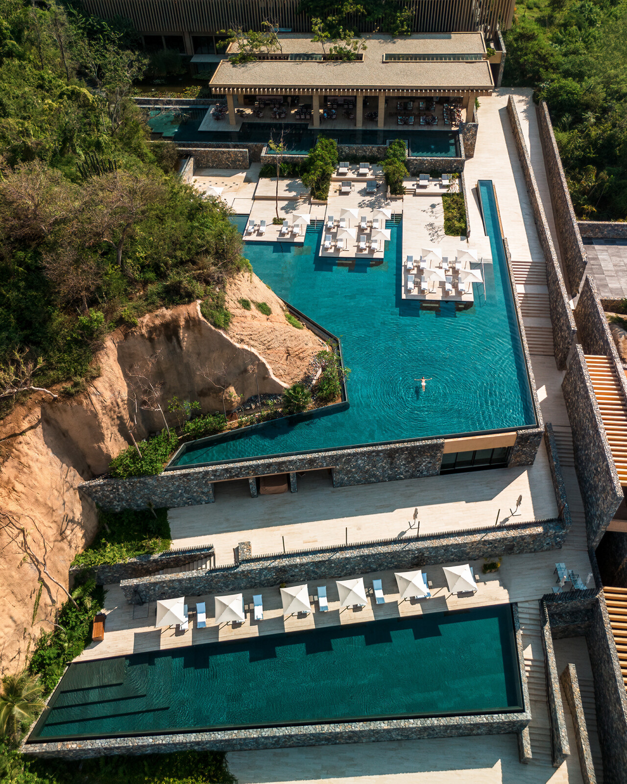 Four Seasons Tamarindo resort's cascading infinity pools on different levels. Shown on a bright sunny day with one swimmer in the top pool. White umbrellas and sun loungers line the pool's edge. Next to a rock formation and green plants.