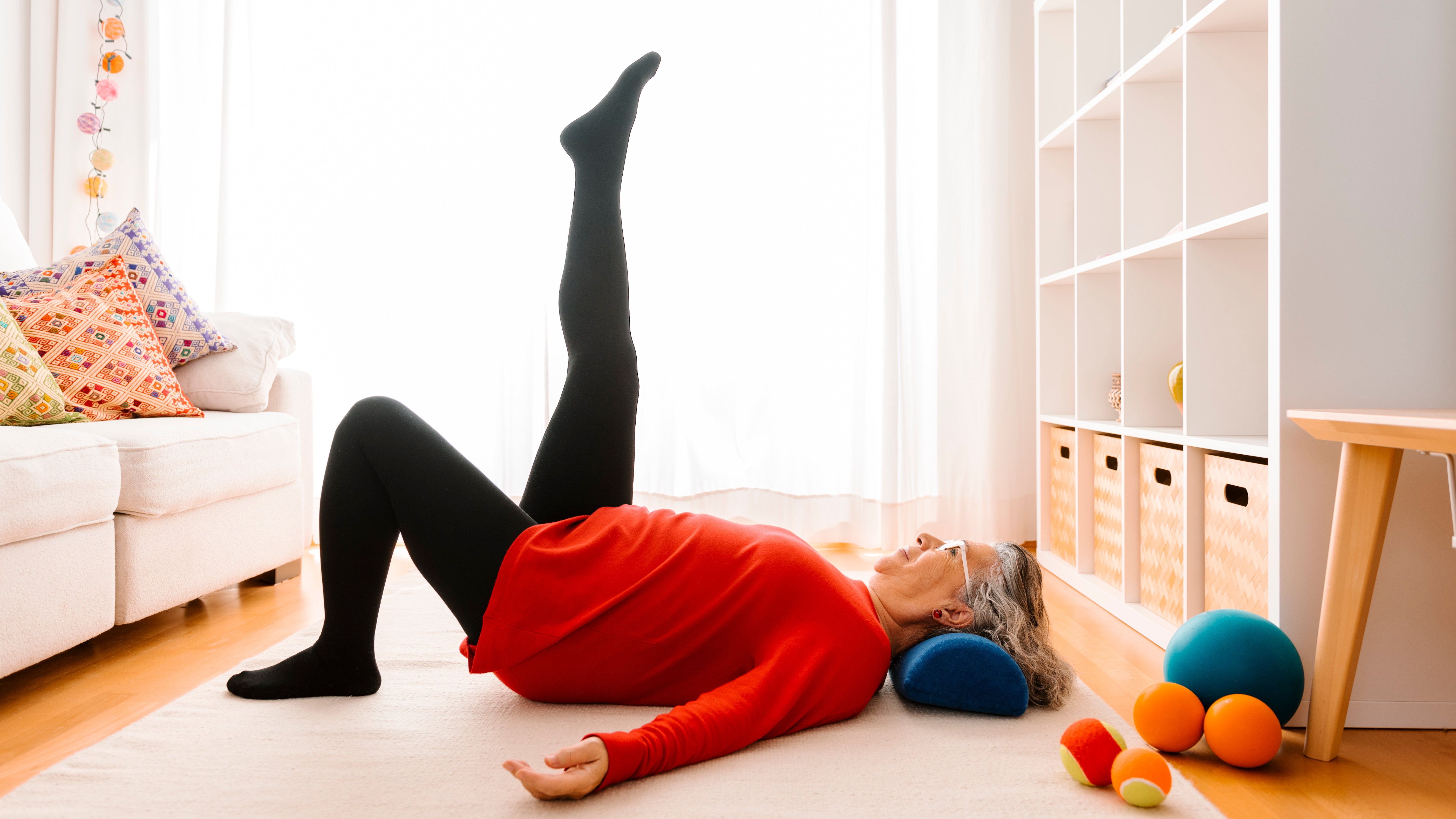 A woman practices Pilates at home. She is on her back, with a cushion under her head, one foot planted on the floor and the other extended toward the ceiling.