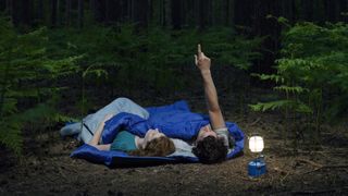 Couple lying in sleeping bags in forest with illuminated lantern, man pointing upwards