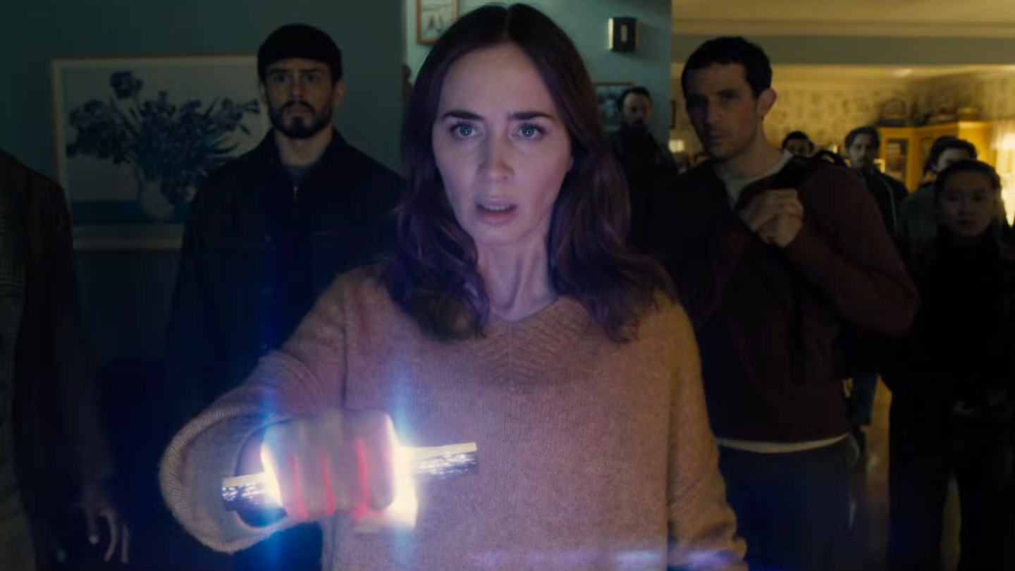 A woman in a sweater (actress Emily Blunt) holds up a glow stick in her fist, as others (including actor Josh O'Connor, right) stand behind her in a living room, in a still from 'Disclosure Day.'