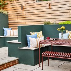 A garden with HAY's Palissade outdoor dining table and matching bench in red next to built-in bench seating painted dark green