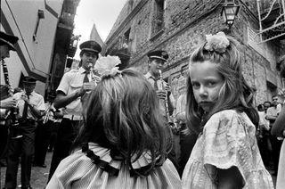 Black and white photograph of two young girls watching an orchestra perform on the street. One of the girls has her head turned to look at the camera