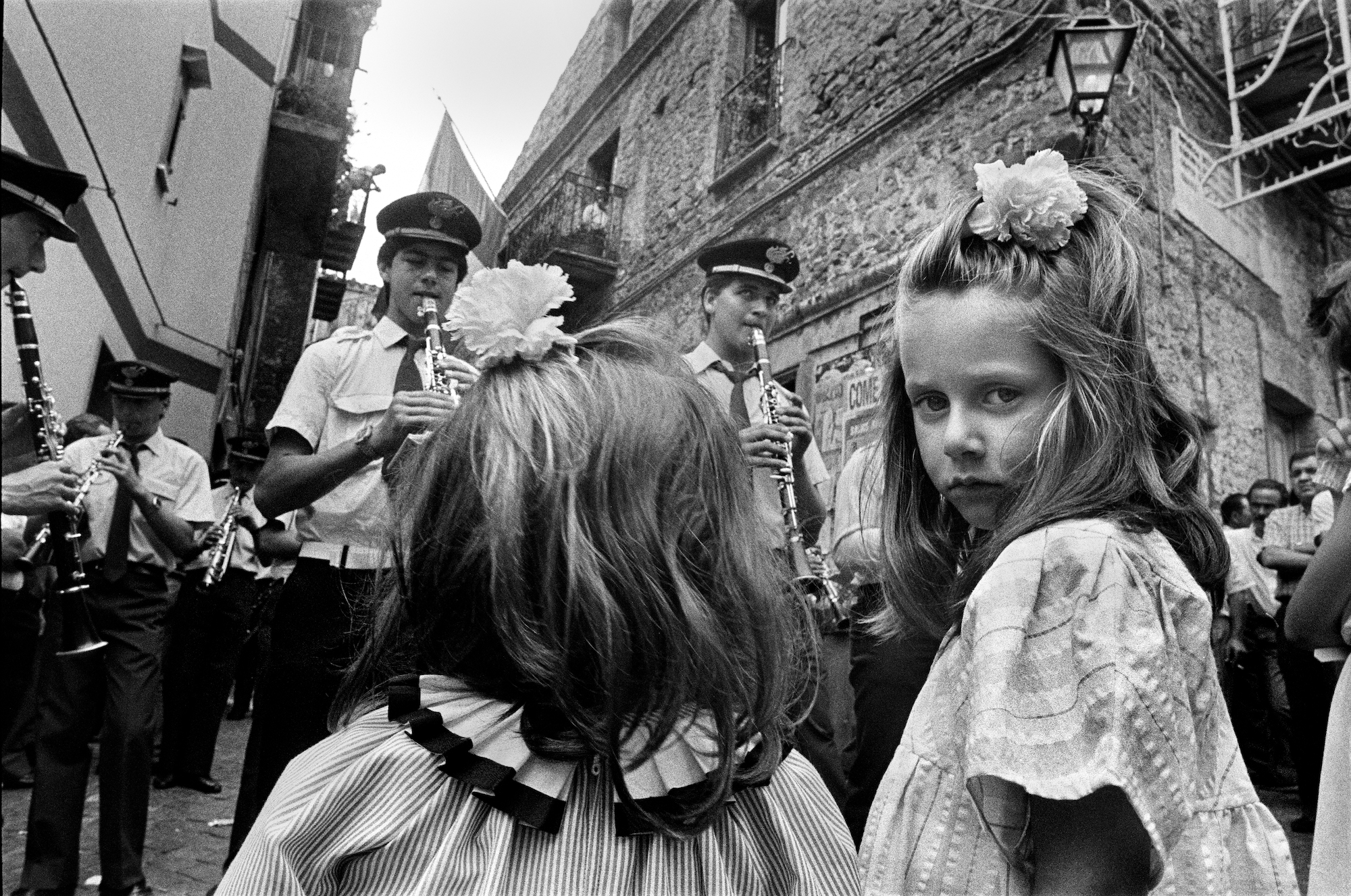 Black and white photo of two young girls watching an orchestra perform on the street. One of the girls turned her head to look at the camera