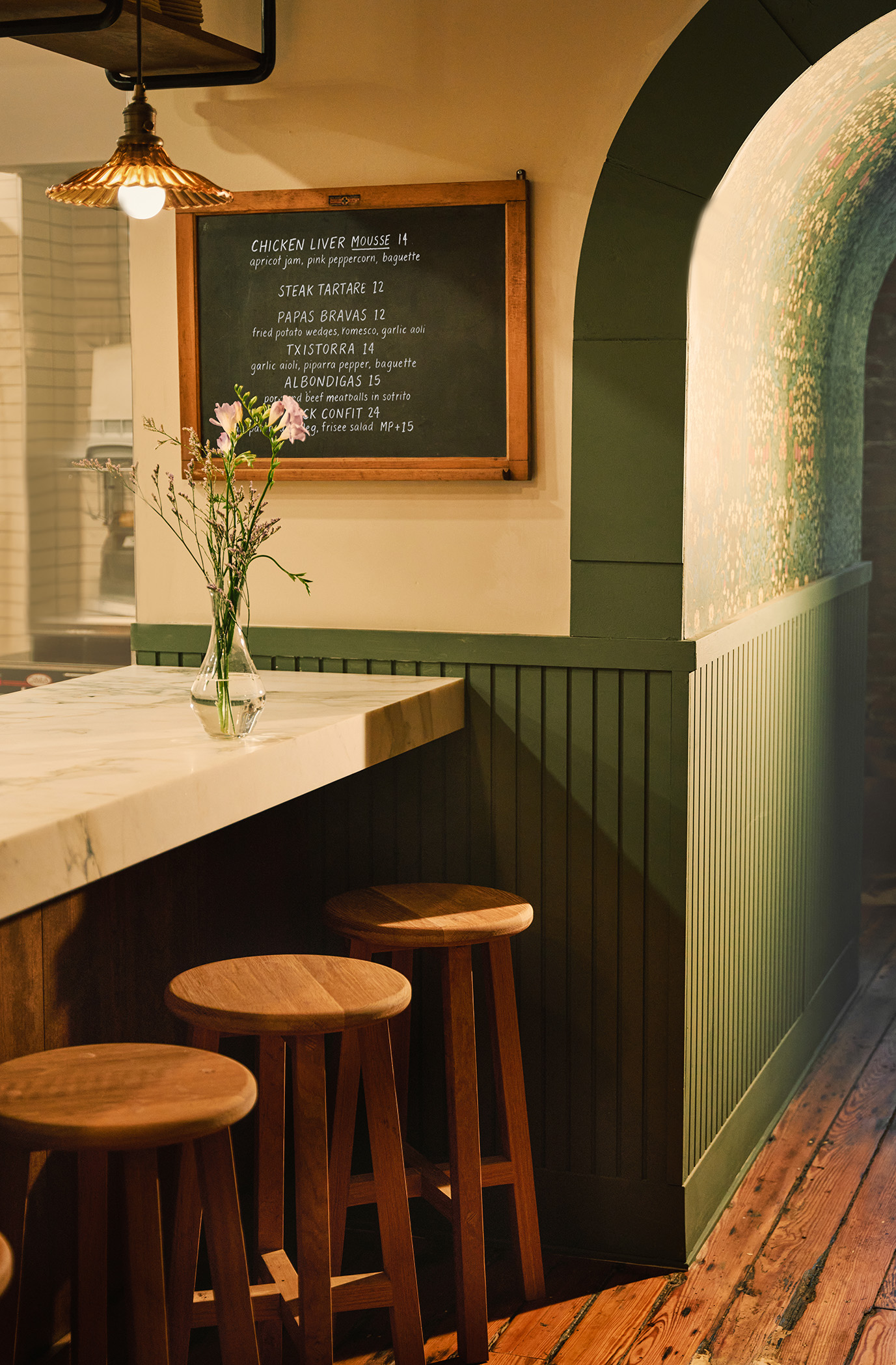 Interior shots of a brick, wood, and marble-clad cafe, bakery, and butcher, decorated with modern rustix furnishings and floral wallpaper.