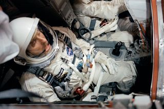a man in a white spacesuit with a glass-domed helmet sits in a cramped spacecraft cockpit