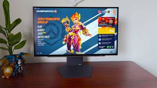 LG UltraGear 27GX790A-B monitor on woodgrain desk next to plant, Pharah figure, Samus toy, and Squishville Bigfoot with main menu on screen featuring Juno.